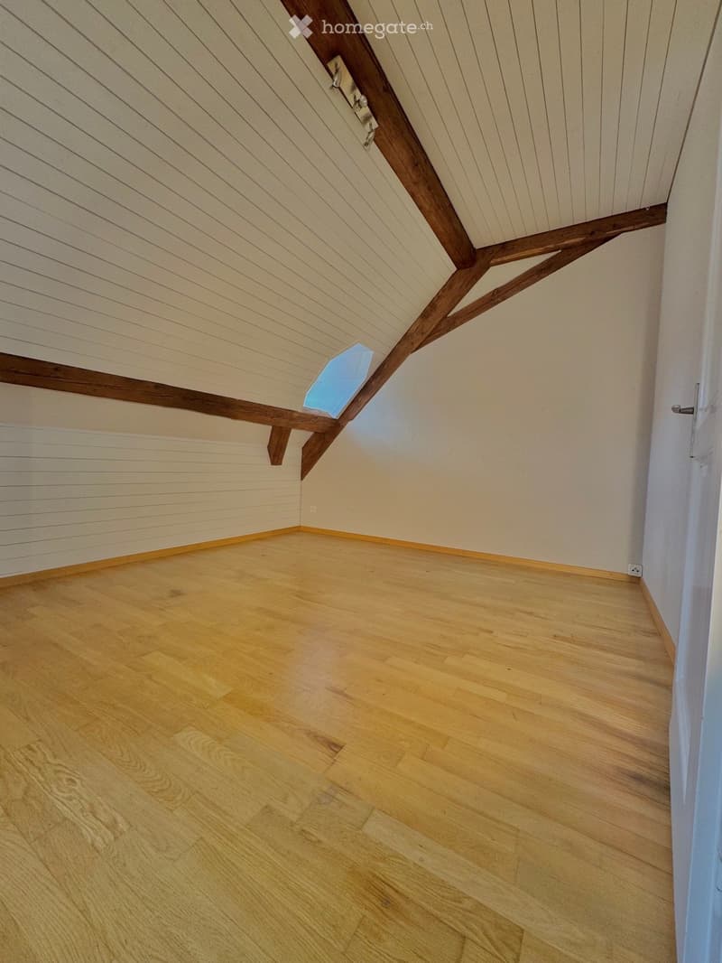 Empty attic room with wooden floor, white walls, and exposed wooden beams on the ceiling. Features a skylight window and a door.