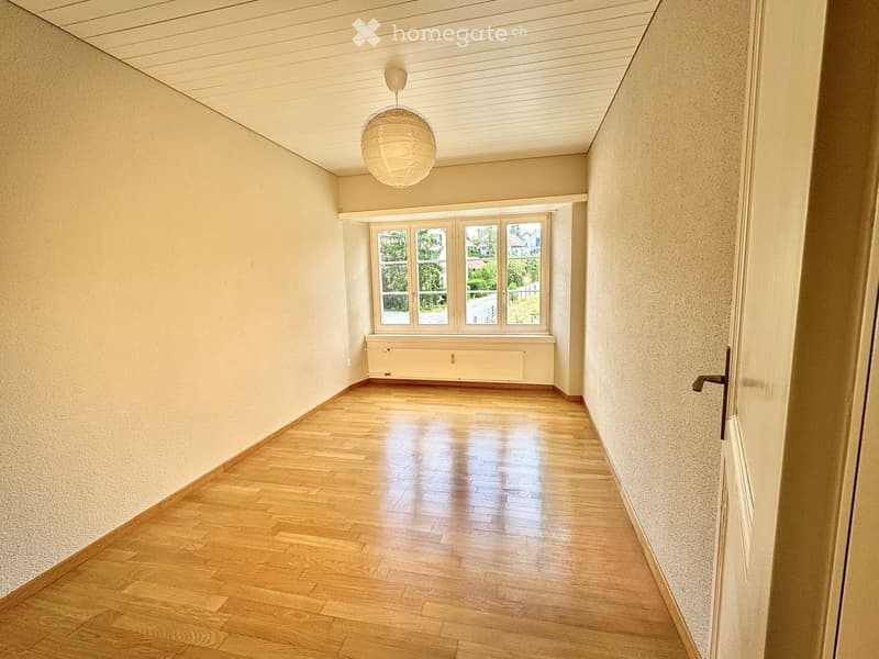 Empty hallway with wooden flooring, light-colored walls, and a large window at the end. A single hanging light fixture is visible.