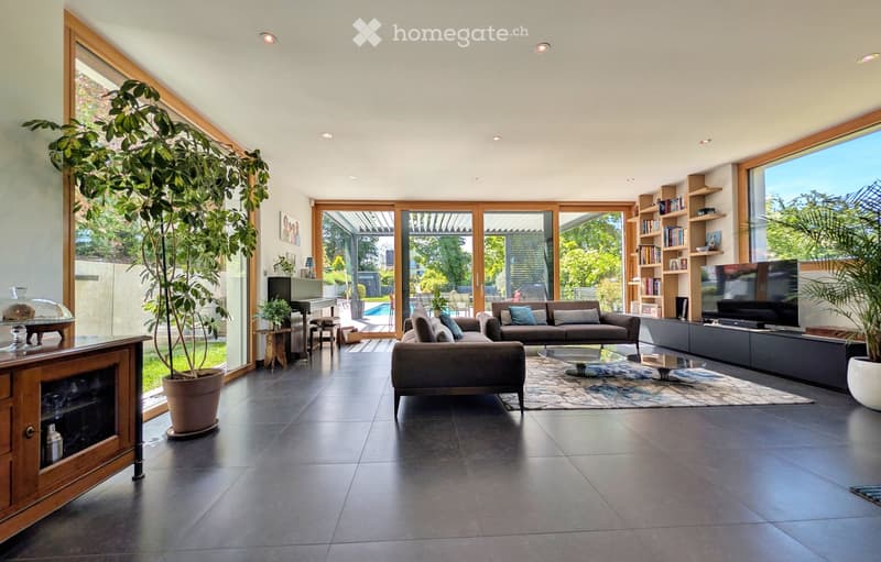 Modern living room with large windows, dark tiled floor, grey sofa, coffee table, bookshelf, piano, and potted plants.