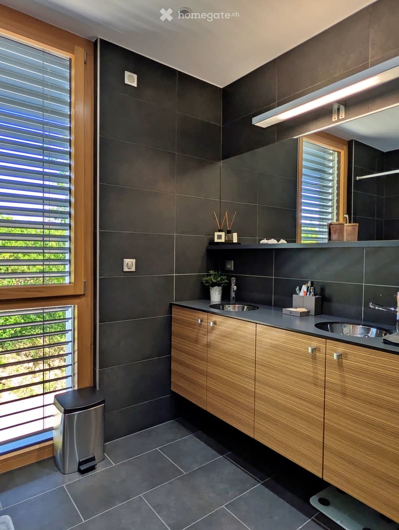 Modern bathroom with dark grey tiles, wooden cabinets, double sink, and large windows with blinds.