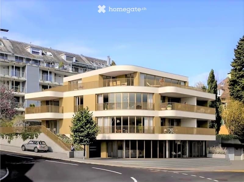 Modern multi-story residential building with large windows, balconies, and contemporary design. The building features a combination of beige and brown tones, with a sleek, curved facade and multiple levels.