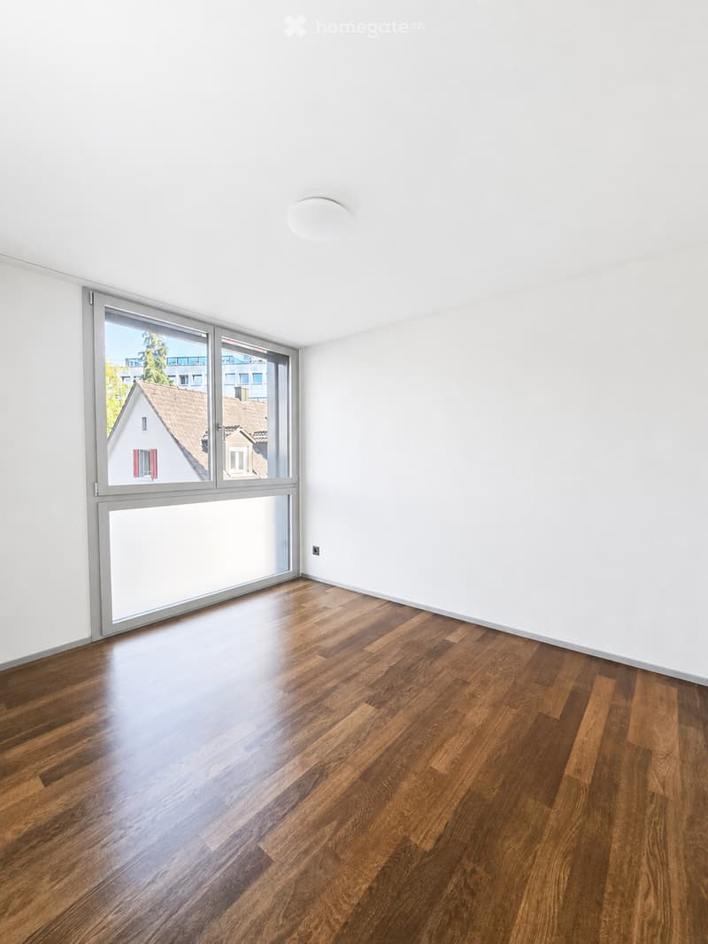 Empty room with wooden flooring and large windows showing a view of neighboring buildings.