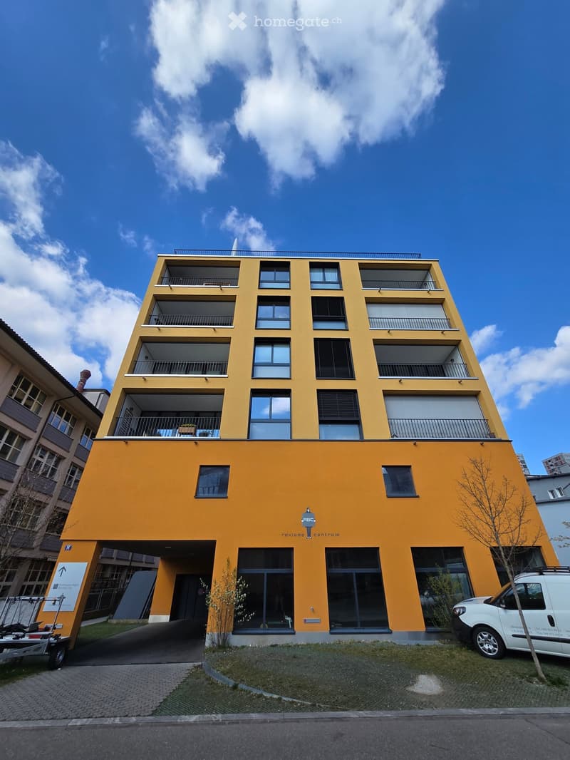 Modern yellow apartment building with multiple balconies and large windows, featuring a parking area and signage for 'Reklame Centrale'.