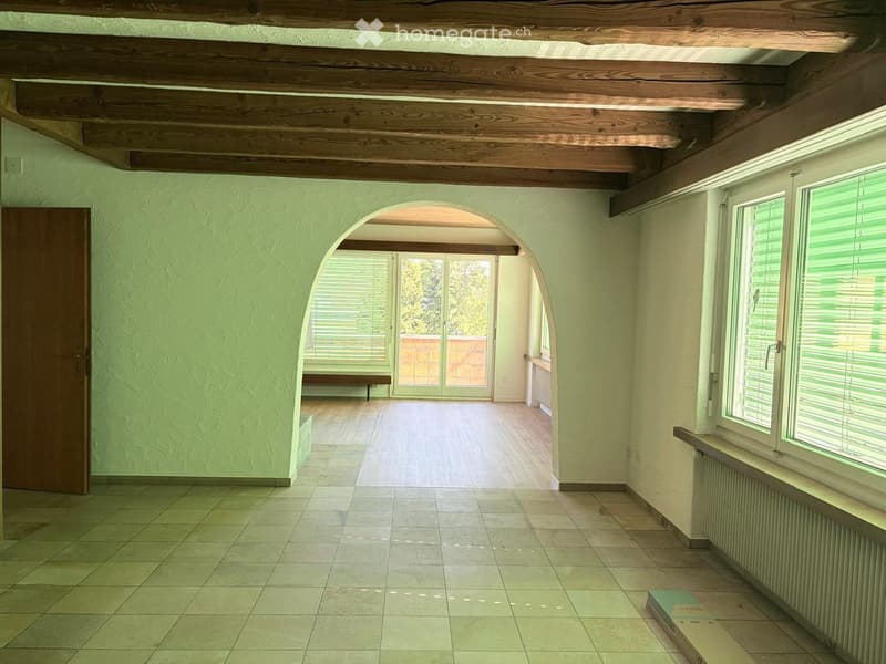 Empty room with tiled floor, wooden ceiling beams, arched doorway leading to another space, and windows with blinds.