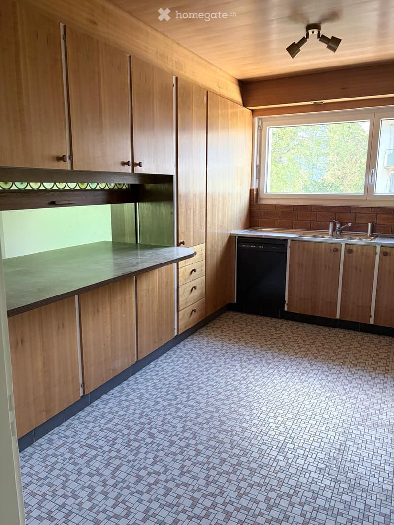 Empty kitchen with wooden cabinets, tiled floor, and a window overlooking greenery.