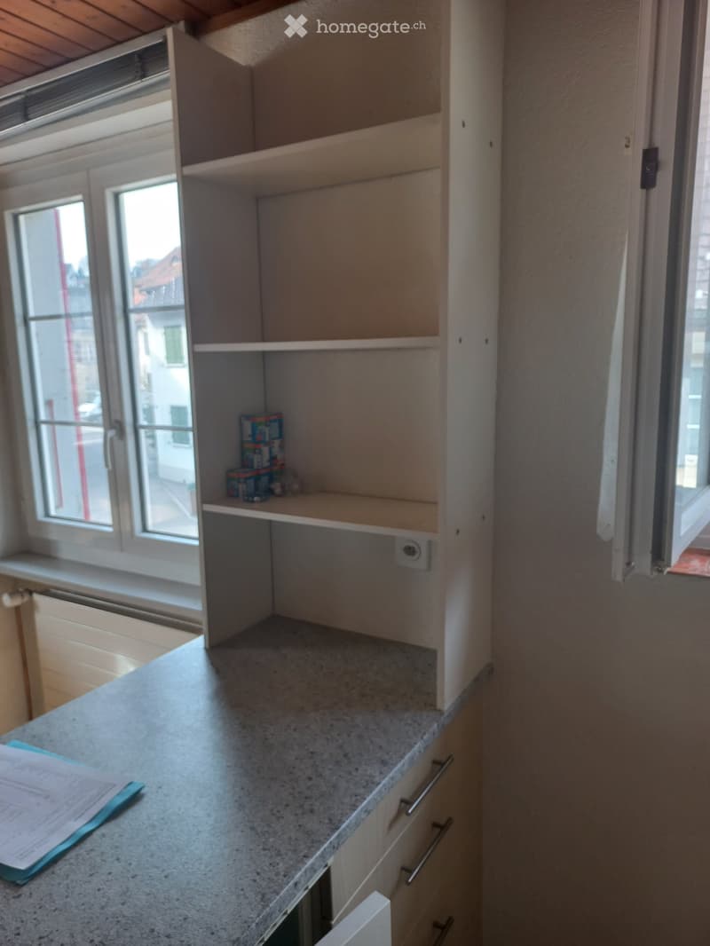 Corner shelf with books and boxes, gray countertop, drawers, window sill, windows, radiator, paper on the counter