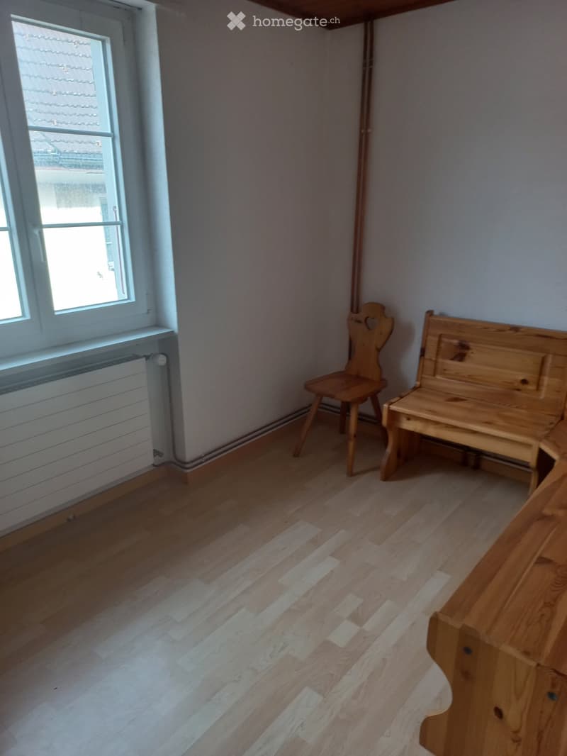 room with white walls, wooden floor, two wooden benches, one wooden chair, a window with white frame, a radiator next to the window
