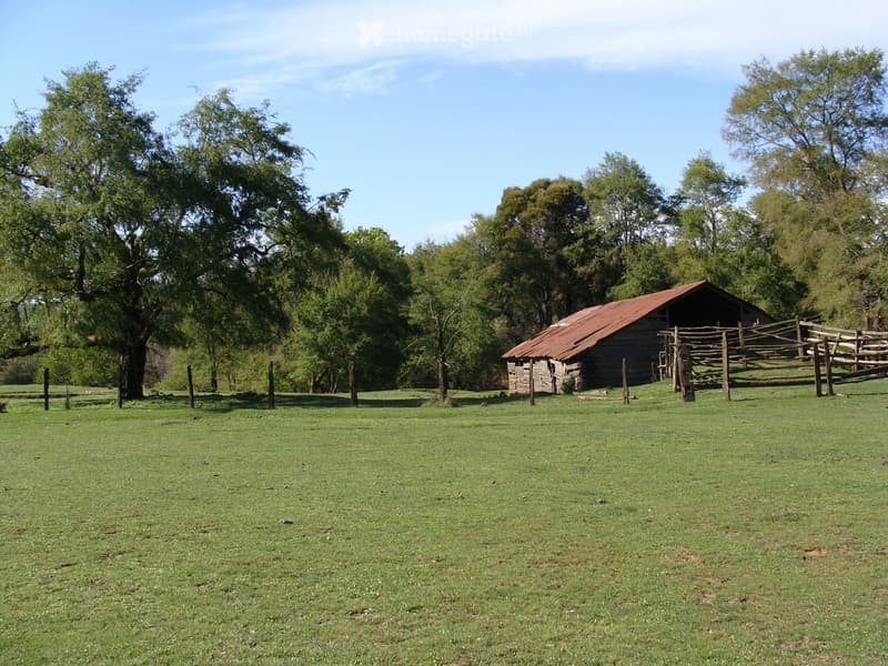 A grassy field with a wooden fence and a small wooden barn with a red metal roof, surrounded by trees.
