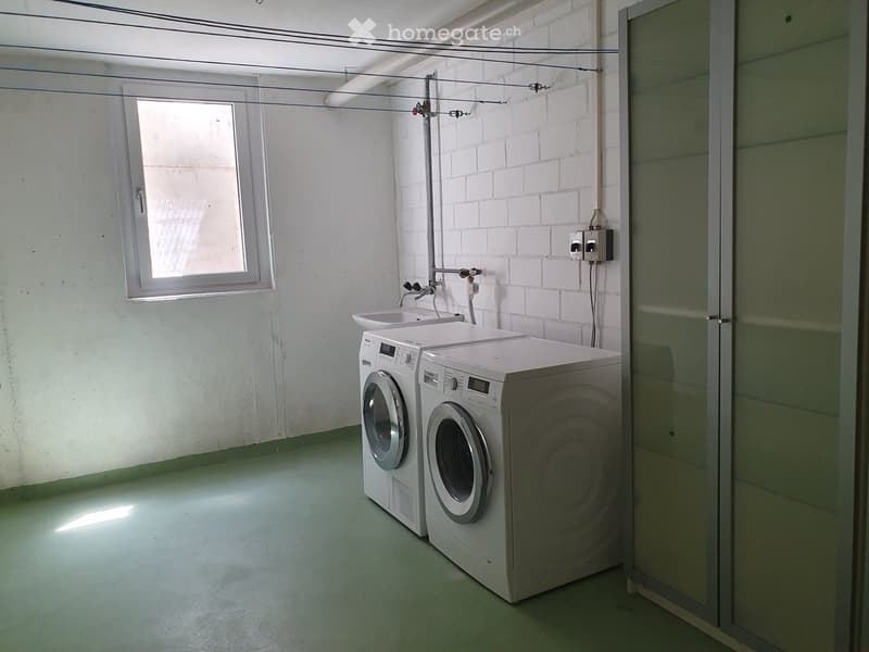 A small, unfurnished utility room with white brick walls, a green floor, and a window. Contains a washing machine and a sink.