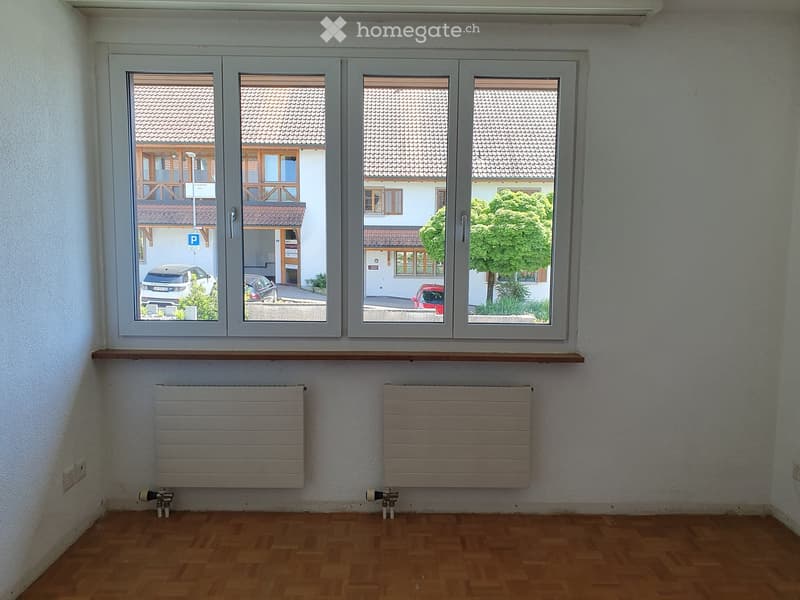 Empty room with wooden flooring, white walls, large windows showing a view of neighboring houses and parked cars, and two radiators below the windows.