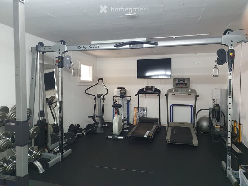 Well-equipped home gym with a treadmill, elliptical machine, weight machine, dumbbells, a large exercise ball, and a mounted TV screen.