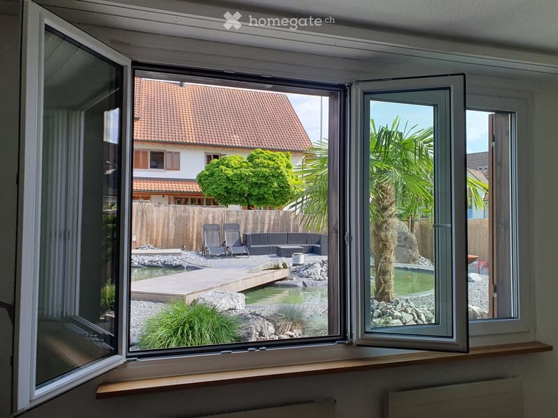 View from a room showing a modern garden with a small pond, wooden deck, lounge chairs, and palm trees.