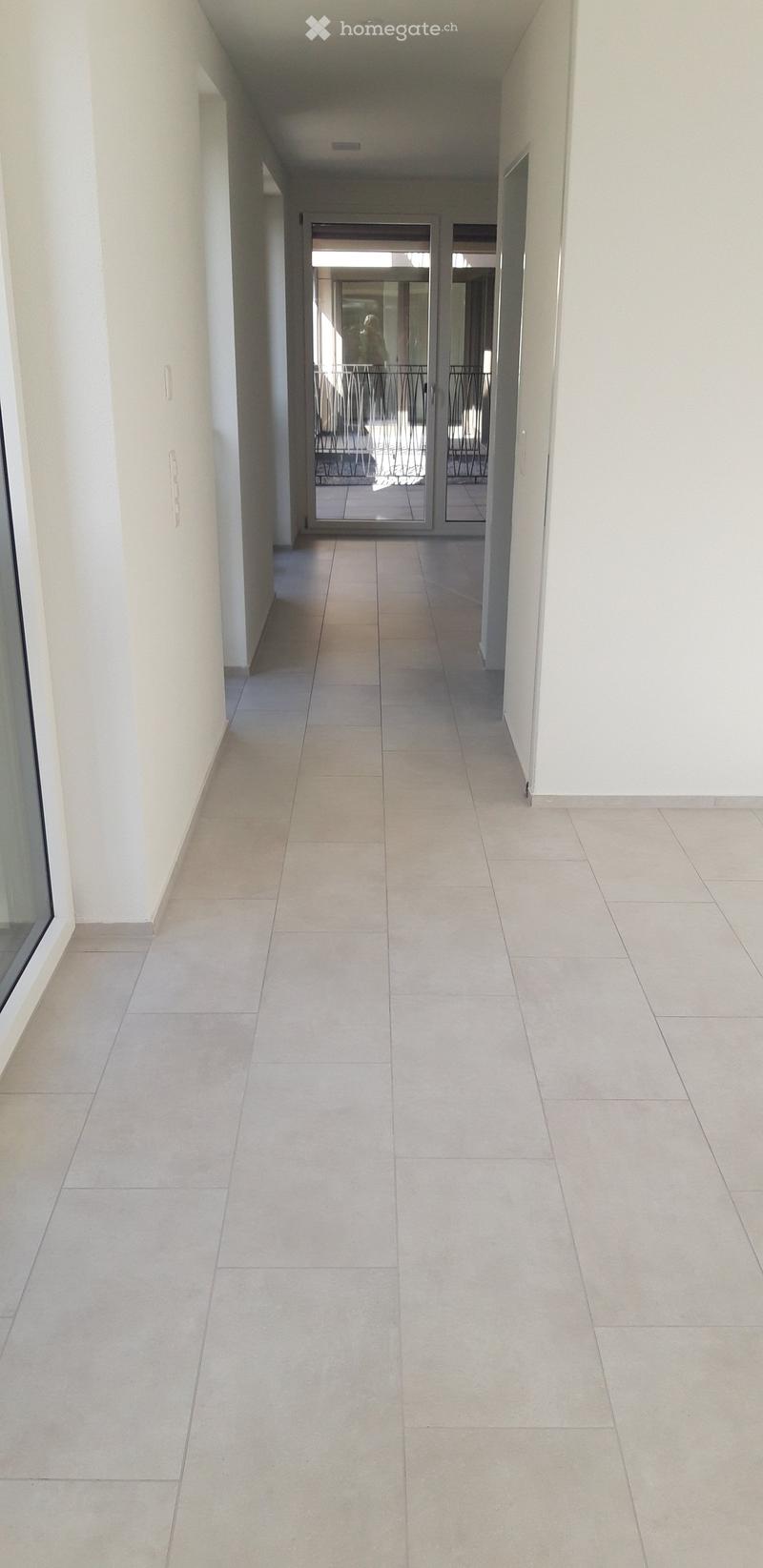 Clean, modern hallway with tiled flooring and white walls, leading to a glass door opening onto a balcony area.