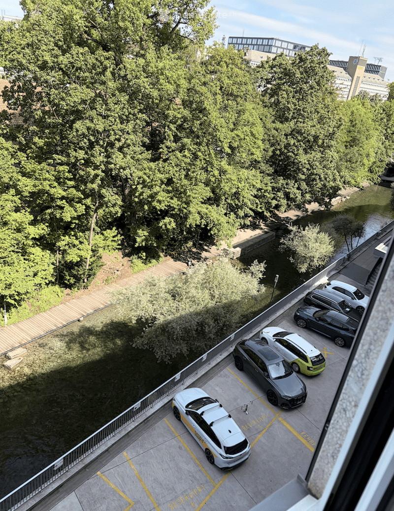 Aerial view of a parking lot with several cars parked, surrounded by greenery and trees, with a wooden walkway and a body of water nearby.