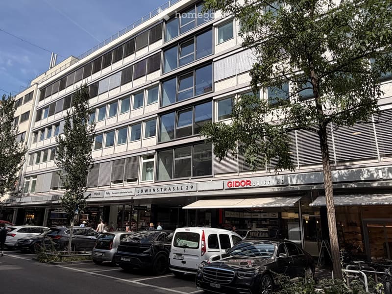 Modern multi-story building with large windows and a storefront labeled "GIDOR COIFFURE" on the ground floor, surrounded by parked cars and trees.
