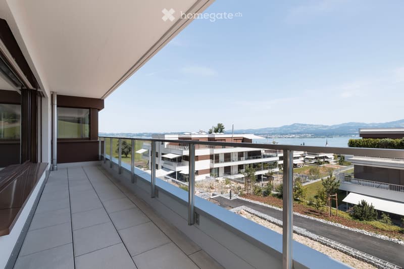 Modern balcony with glass railing, tiled floor, overlooking a waterfront with modern buildings and distant mountains.