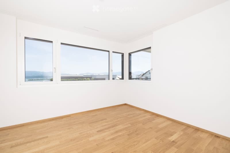 Empty room with white walls and wooden flooring, featuring large windows with scenic views.