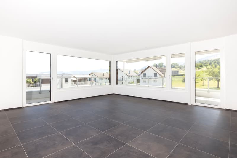 Empty room with dark tiled floor, white walls, large windows, and sliding glass doors leading to a balcony with a scenic view of houses and mountains.