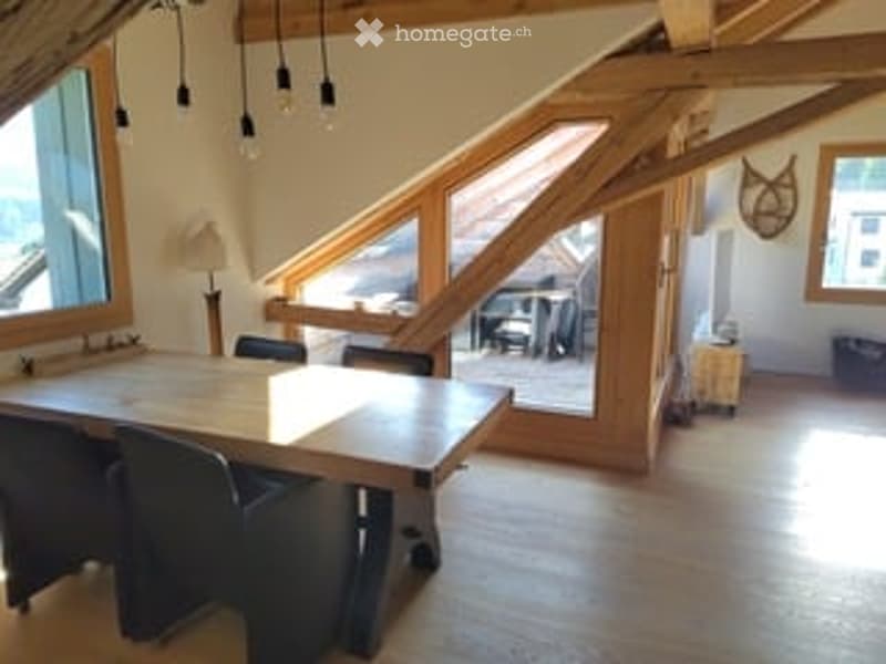Bright, airy room with wooden beams and large windows, featuring a dining table with chairs, a floor lamp, and a small stool near the window.