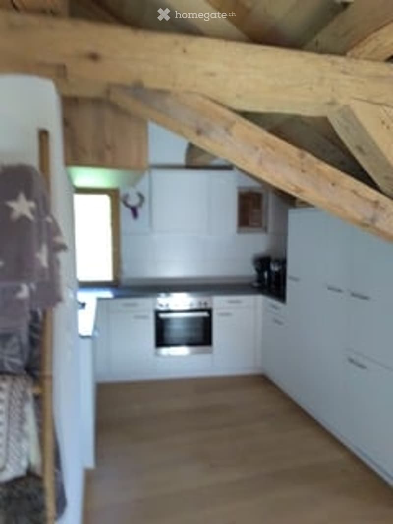 Bright kitchen with white cabinets, wooden ceiling beams, and a wooden floor. Features an oven, coffee maker, and a window letting in natural light.