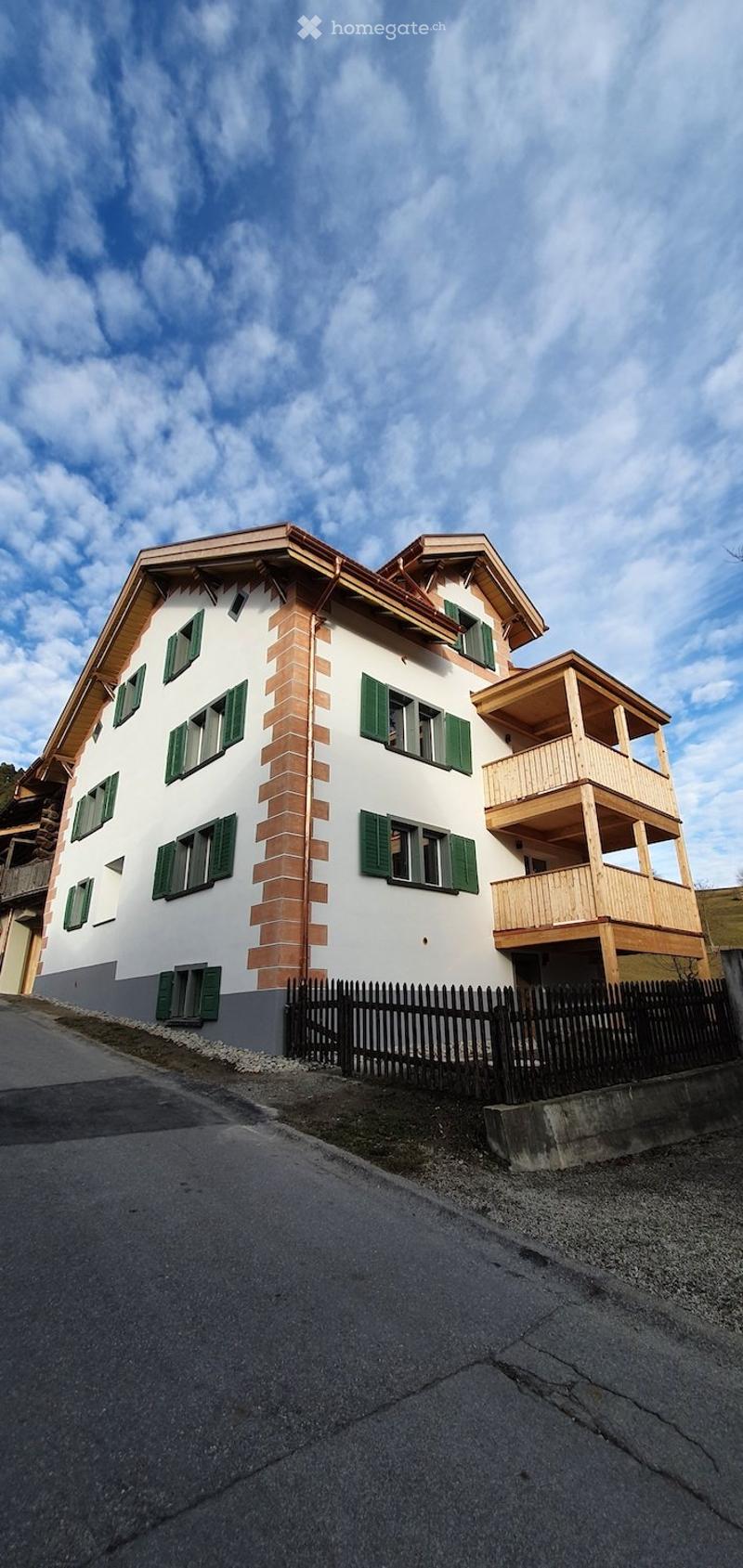 Two-story house with white walls, green shutters, and a wooden balcony on the upper level.