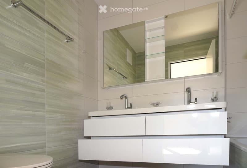 Modern bathroom with a white vanity cabinet, double sink, sleek mirror, and glass shelves. The walls are tiled in a light, marble-like pattern.