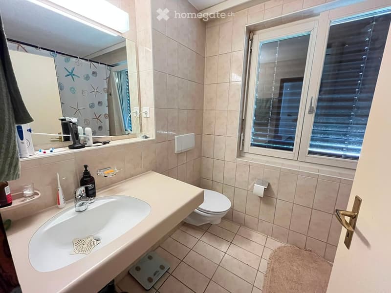 Bathroom with beige tiles, a large sink, a toilet, a mirror, and a window with blinds. Personal care items are placed on the sink counter.