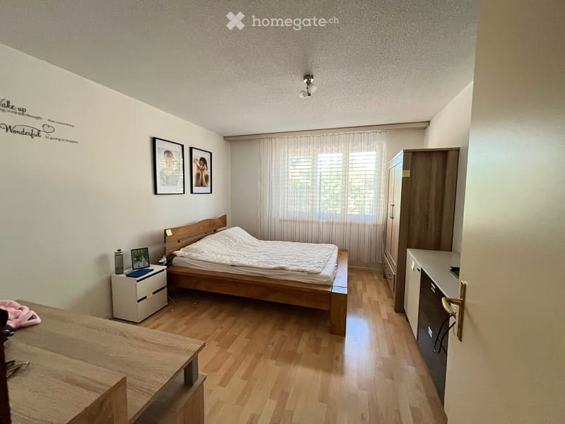 A well-lit bedroom with wooden flooring, a bed with white bedding, a nightstand with a lamp, a small table, a wardrobe, and framed pictures on the wall.