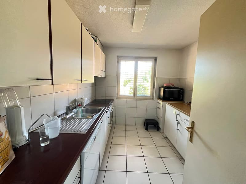 Modern kitchen with white tiled walls and floors, white cabinets, a dark brown countertop, a sink with a faucet, a microwave, a blender, and a small stool near the window.