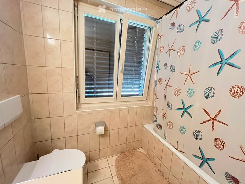 Bathroom with tiled walls and floor, a toilet, a shower curtain with sea-themed designs, a toilet paper holder, and a bath mat.
