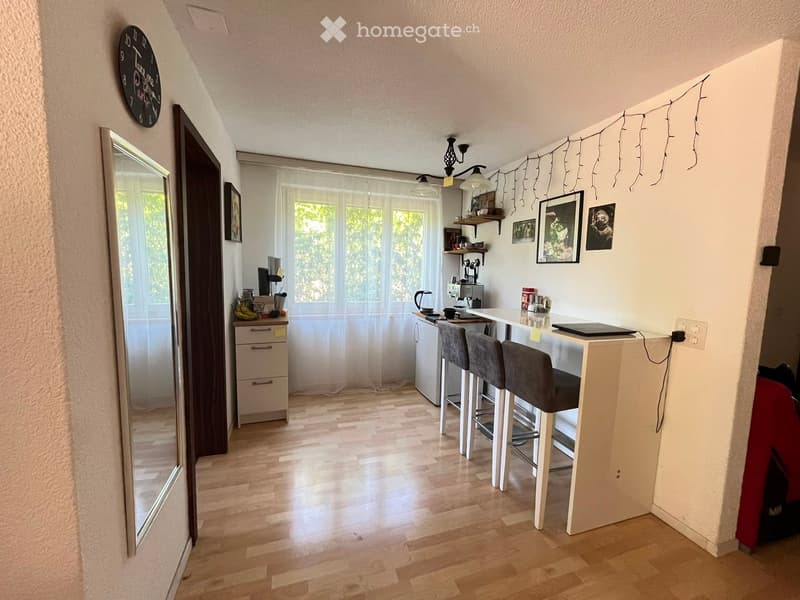 Modern kitchen with wooden flooring, white cabinetry, and a small bar area with two stools. Features a refrigerator, coffee maker, and a laptop on the counter.