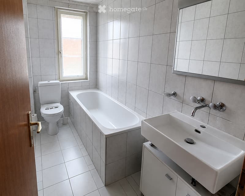 Modern bathroom with white tiles, a toilet, a bathtub, a sink with a modern faucet, a mirror, and a cabinet under the sink.