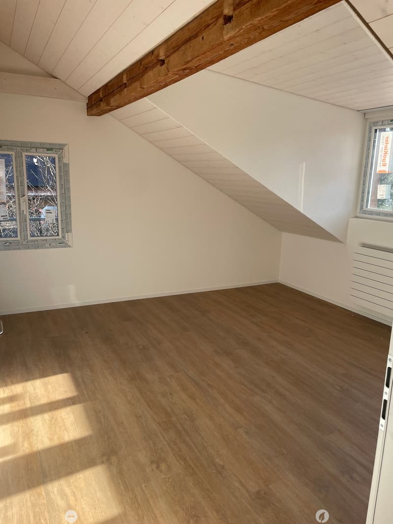 Unfurnished Room. Empty attic room with wooden flooring, white walls, and exposed wooden beams on the ceiling. Two windows with frosted glass panels are visible.