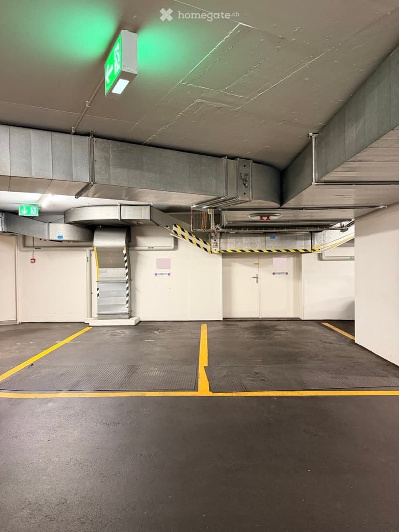Empty underground parking area with marked parking spaces, white walls, and industrial ceiling with exposed pipes and lighting.