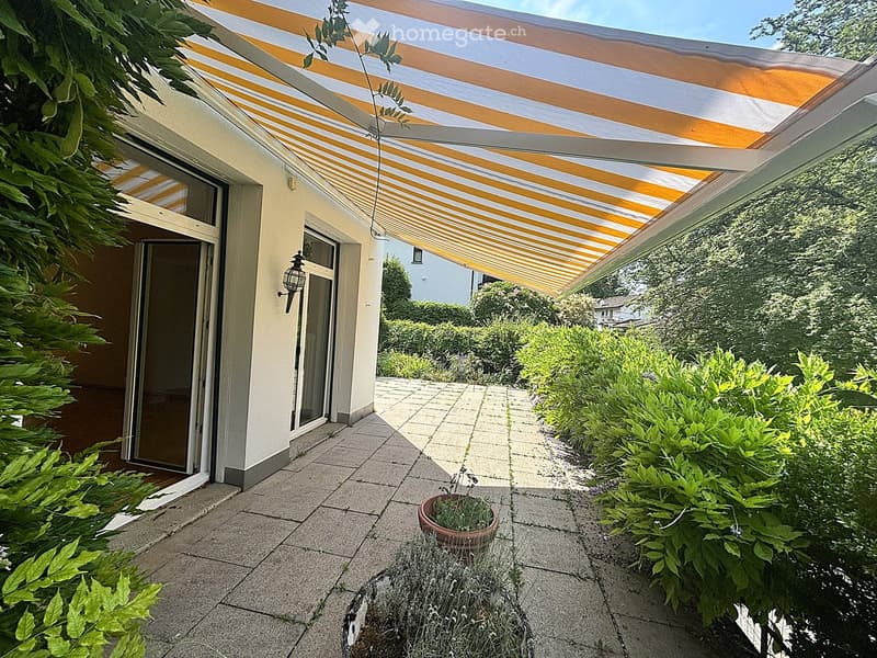 Outdoor patio area with a striped awning, tiled floor, potted plant, and surrounding greenery.