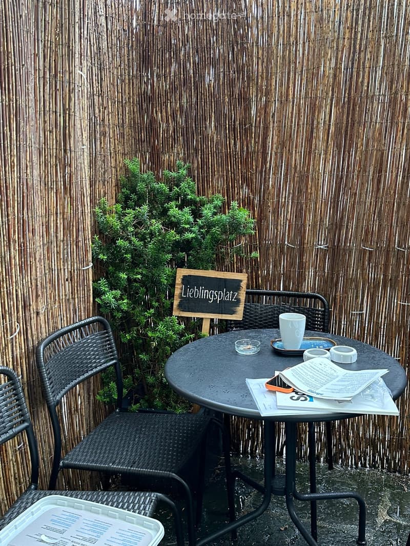 Outdoor seating area with a round black metal table, two wicker chairs, a cup, and a sign that reads "Lieblingsplatz" in German.