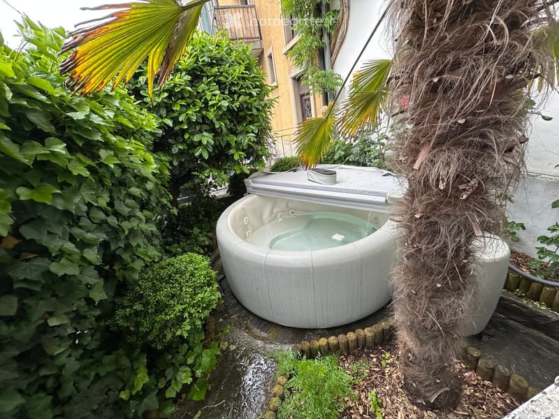 A small, round hot tub surrounded by lush greenery, including palm leaves and ivy, with a wooden fence and a building in the background.