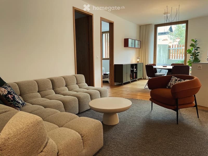 Modern living room with a sectional sofa, coffee table, armchair, dining area with table and chairs, and a cabinet. The space features large windows and wooden flooring.