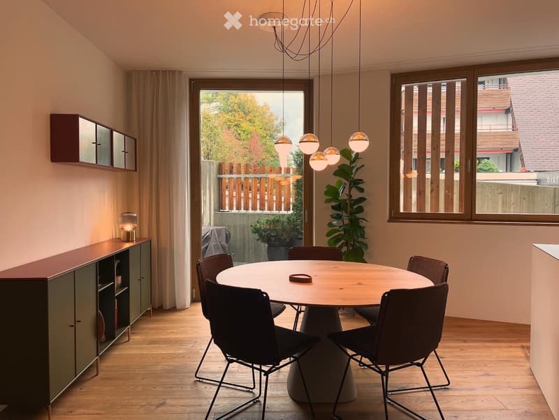 Modern dining area with a round wooden table, six chairs, pendant lights, a tall plant, and a cabinet with a mirror on the wall.