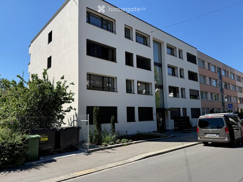 Modern multi-story residential building with white facade, large windows, and black shutters. Features a small garden area with greenery and trash bins, and a parked van on the street.