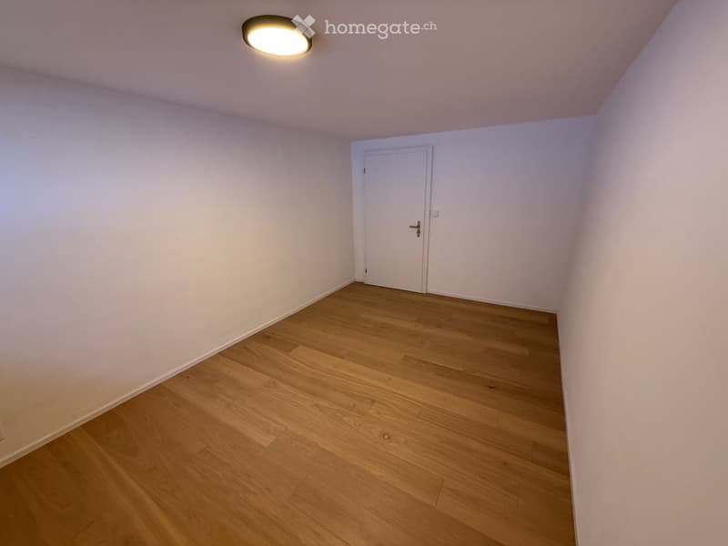 Empty room with light wooden flooring, white walls, and a single white door. A round ceiling light fixture is visible.