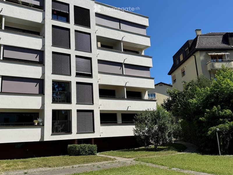 Modern multi-story apartment building with balconies, large windows, and greenery in front.