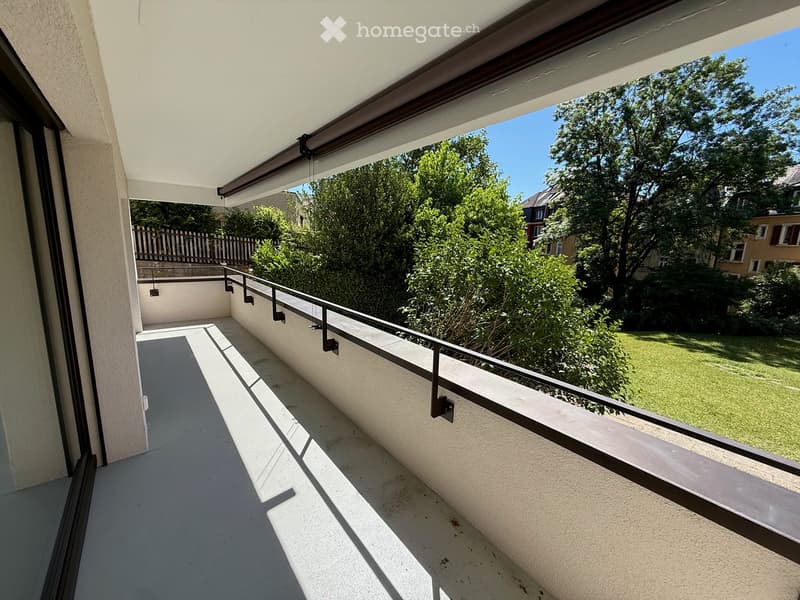 Balcony with modern design, white walls, metal railing, and a retractable awning, overlooking a green garden area.