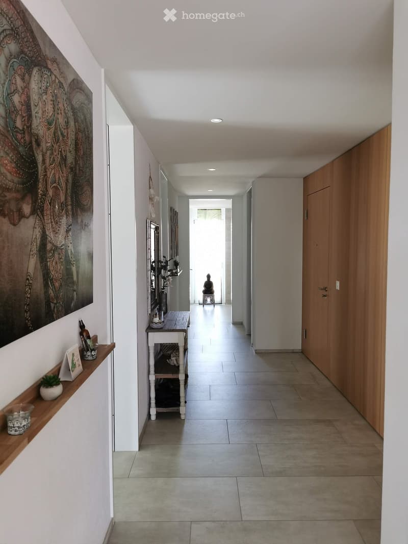 Modern hallway with white walls, tiled floor, wooden shelves, and artwork. Features a small table with decorative items and a small plant on a shelf.