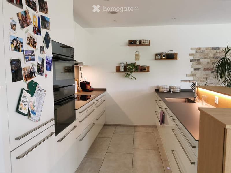 Modern kitchen with white cabinets, stainless steel appliances, and a wooden island. Features include a refrigerator with photos, an oven, a sink with a modern faucet, and decorative shelves with plants and jars.
