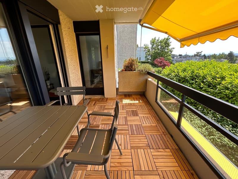 Balcony with wooden decking, table, chairs, and a yellow awning, overlooking greenery and a cityscape.