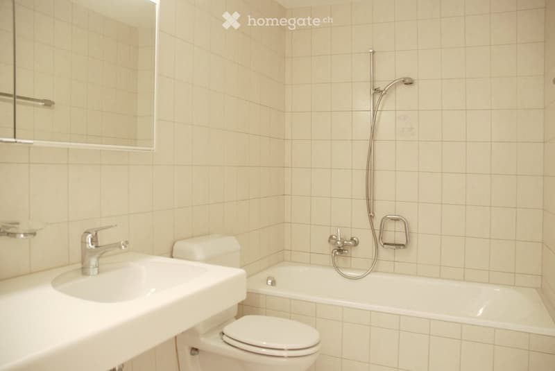 Bathroom with beige tiled walls, a white sink with a chrome faucet, a toilet, a bathtub with a showerhead, and a mirror above the sink.