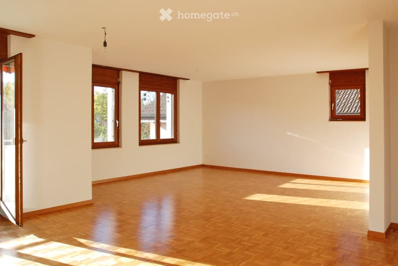 Empty room with wooden flooring, white walls, and multiple windows with wooden frames. Natural light streams in, creating shadows on the floor.