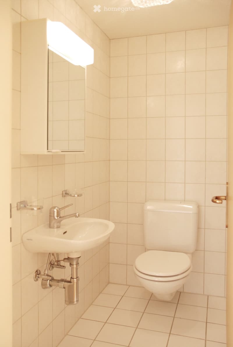 A small bathroom with a white sink, chrome faucet, and a white toilet. The walls and floor are tiled in light beige.