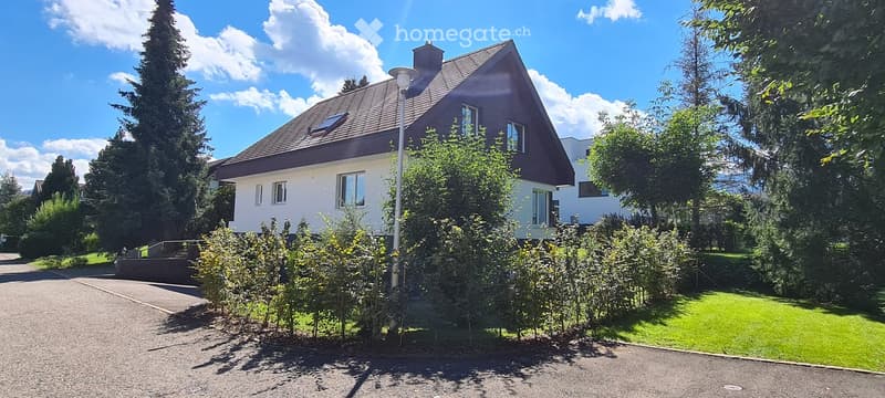 Two-story house with a dark brown roof and white walls, surrounded by greenery and a well-maintained lawn.
